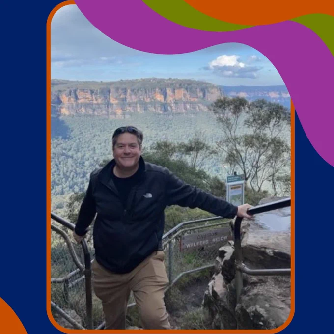 Tom Mehen smiles at the camera while holding onto metal rails. Behind him is an expansive view of a canyon.