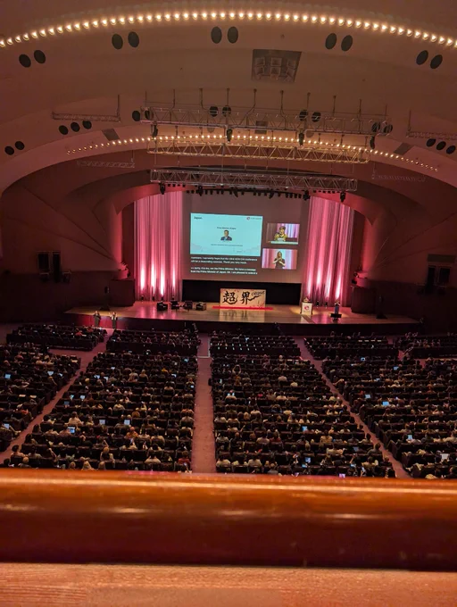 A large auditorium filled with attendees seated in rows, facing a brightly lit stage with pink curtains. A presentation is being projected on a large screen, showing a message from Japanese Prime Minister Shigeru Ishiba welcoming the CHI2025 community. The CHI2025 logo and Japanese calligraphy are displayed on stage.
