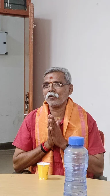 Prakash, an older man with gray hair and a mustache, is sitting at a table. He is wearing glasses, a red shirt, and an orange shawl draped over his shoulders. He has a red bindi on his forehead and his hands are joined together in a gesture of greeting or prayer. On the table in front of him, there is a yellow paper cup and a blue-capped plastic water bottle. A wooden door is partially visible behind him., In list, Window Ask more