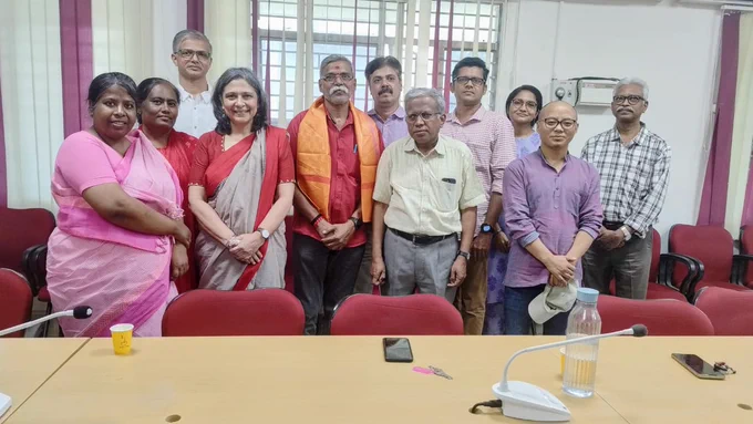 A group of eleven people is standing together in a room. They are positioned in two rows, with some seated and others standing behind them. The individuals are of diverse ages and appear to be of South Asian descent. The women are wearing traditional Indian attire, such as sarees, while the men are dressed in casual shirts and trousers. Prakash, a man in the front row, is wearing an orange shawl over his shoulders. The room has red chairs and a table in front of the group, on which there are a microphone, a water bottle, a small yellow cup, a mobile phone, and a set of keys. The background features windows with bars and curtains., In list
