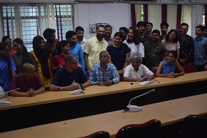 A group of people is gathered in a conference room. There are about 30 individuals, with a mix of men and women, standing and sitting around a long, curved conference table. The front row consists of five older men sitting at the table, with microphones in front of them. Behind them, a larger group of younger individuals stands, smiling by seeing towards prakash. The room has maroon and white vertical blinds on the windows, ceiling fans, and air conditioning units. There are also ceiling lights and a projector mounted on the ceiling. On the table, there is a green water bottle and a notebook with a pen.