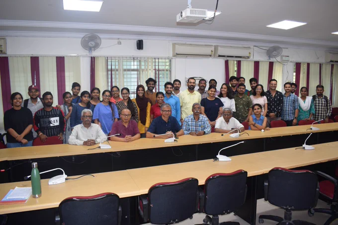 A group of people gathered in a room for Prakash's retirement ceremony. Prakash, the man in a blue and white checkered shirt, is cutting a cake placed on a table to celebrate his retirement. Around him, several people are standing, watching, and smiling, with some clapping. A woman in a red shirt stands next to the table, while another woman captures the moment with her phone. The room features a television mounted on the wall, a cabinet, and a blue office chair, creating a casual and cheerful atmosphere for the celebration.