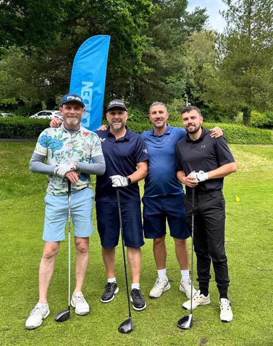 Photograph taken at our 2024 Charity Golf Day. A team of four golfers are stood in a line on the golf course, three of them are each holding a golf club. They are all smiling at the camera. Behind them is a blue feather flag and in the background are several trees.