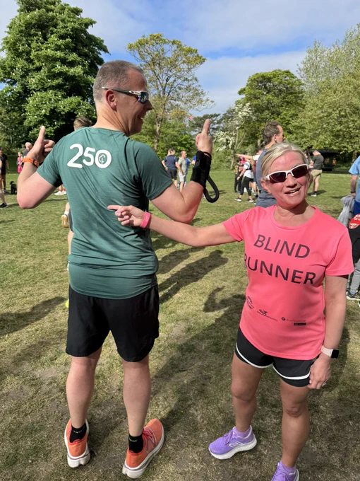A man and a woman are standing on a grassy area with other people in the background. The man is wearing a green shirt with the number "250" on the back, black shorts, and orange running shoes. He is pointing his thumbs towards himself and smiling. The woman is wearing a bright pink shirt with the words "BLIND RUNNER" printed on it, black shorts, and purple running shoes. She is pointing at the man and smiling. Both are wearing sunglasses, and the woman has a smartwatch on her wrist. The scene is set in a park with trees and a clear blue sky.