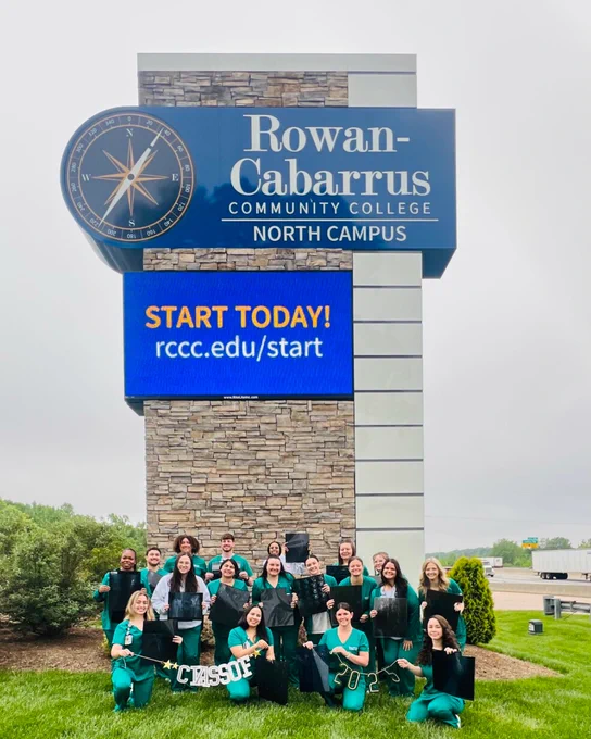 A group of graduates in green scrubs pose in front of the Rowan-Cabarrus Community College North Campus sign, holding their caps and celebratory signs that read “Class of 2025.” The digital sign above them displays the message “START TODAY! rccc.edu/start.” The setting is outdoors on a cloudy day with grass and trees in the background.