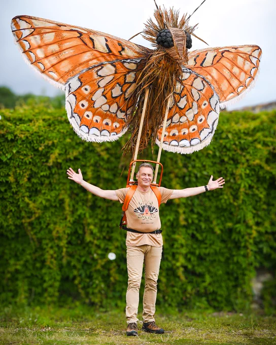 Chris Packham standing in front of a hedge with arms outstretched, wearing a large Marsh Fritillary butterfly puppet on his shoulders