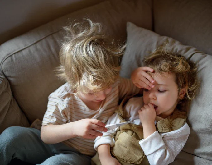 Two children looking after each other on a sofa, one taking the other's temperature with a toy thermometer.