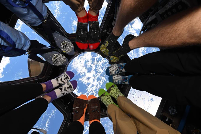A view of astronauts, posed in a circle, wearing colorful socks aboard the International Space Station. The background is a view of the Earth out of the cupola windows.