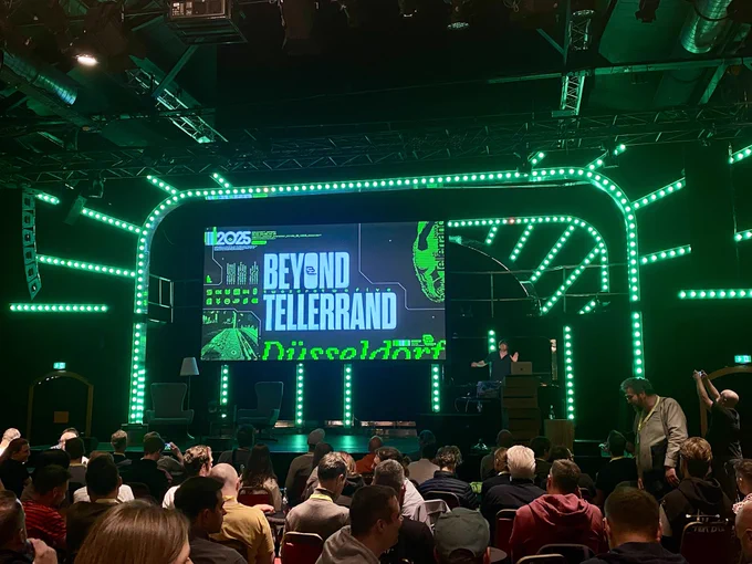 A conference stage featuring a large screen displaying the event title "BEYOND TELLERRAND" in Düsseldorf. The stage is illuminated with green lights, and the audience is seated in front.