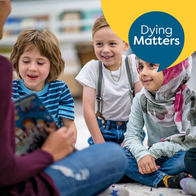 Dying Matters graphic. Photograph of three young children sat together on the floor. An adult is sat in front of them holding an open story book. The children are engaged and smiling with the adult.
