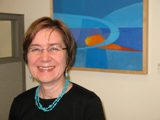 Gosia Fort, a white woman with short brown hair and glasses, standing in front of an organe and blue painting.