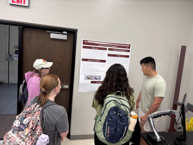 Four individuals are standing in front of a poster titled "The Effects of Cold Temperature on Fall Armyworm Development" at an academic conference. They are engaged in discussing the content displayed.