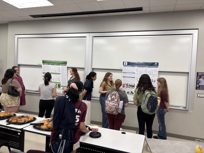 Attendees at a university poster session, engaging in discussions in front of several displayed poster boards in a classroom with whiteboards and refreshments on the tables.
