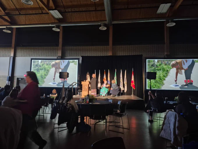 An atrium filled with people and a person behind a podium presenting on stage in front of a row of flags with 2 others seated on stage.