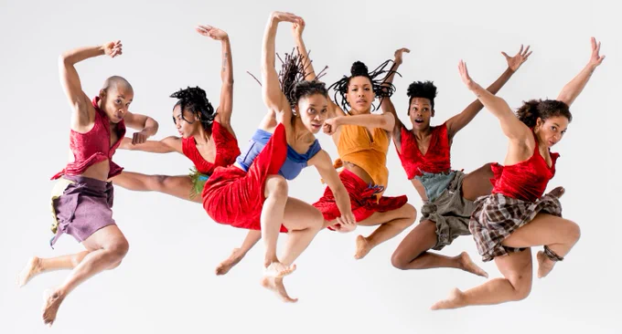 Six dancers in colorful attire perform a dynamic jump against a white background.