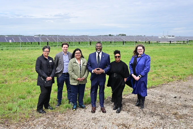 Group of six individuals, including Chicago Mayor Brandon Johnson, First Lady Stacie Johnson, Chief Sustainability Officer Angela Tovar, and Department of Environment staff, standing in front of a large solar panel array in a field, dressed in business-casual attire, smiling towards the camera.