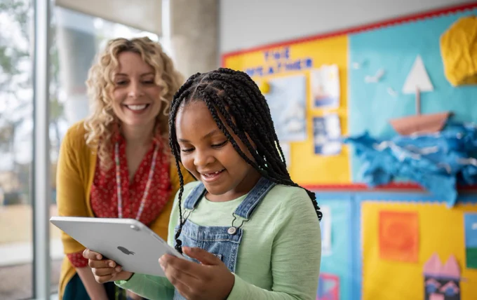 A student in a green shirt and overalls holds iPad, looking at it and smiling. To the right of the student, a teacher in a red shirt and yellow sweater watches as the student uses iPad. They both stand in a classroom in front of yellow and blue bulletin boards.
