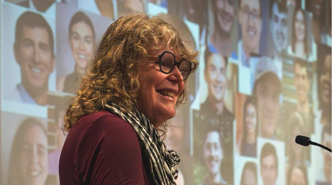 A person with curly hair and glasses smiling while giving a speech at a lectern, with a large background screen displaying multiple faces.
