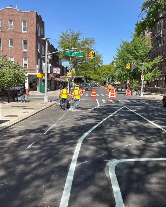Two N Y C D O T workers in reflective yellow safety vests walk along a freshly paved street with new white road markings in place, including a large turn arrow and bike lane lanes. Orange construction barrels and “Do Not Enter” signs block traffic further down the street. The intersection is at 55th Street, surrounded by apartment buildings, shops, and trees on a sunny day. 