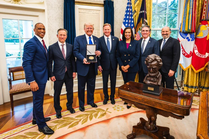 A group of seven people standing in the Oval Office. From left to right: Scott Turner (Secretary of Housing and Urban Development), Josh Harris (Managing Partner of the Commanders), Donald Trump (U.S. President), Roger Goodell (NFL Commissioner), Muriel Bowser (Washington, D.C. Mayor), Doug Burgum (Secretary of the Interior), and Howard Lutnick (Secretary of Commerce). Trump is holding a framed gold coin with the NFL logo on it. 