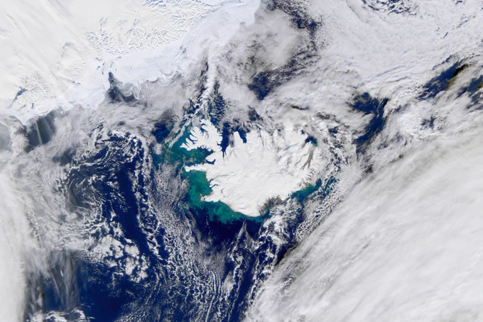 Image Description: An image of Iceland. White clouds cover most of the image. In the middle a white Iceland appears. Around it is a blue ocean with green swirls of phytoplankton.
