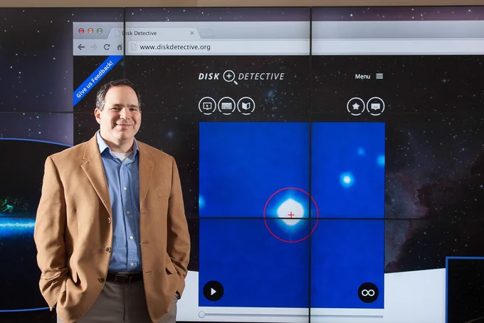NASA astrophysicist Marc Kuchner stands in front of a large display screen showing the Disk Detective website, which aids in studying planet formation. Kuchner, a pioneer for participatory science, is the NASA Citizen Science program officer and has been a principal investigator for various citizen science projects. He is seen wearing a tan blazer and standing with his hands in his pockets. The display on the screen highlights an image from the Disk Detective project. Image credit: David Friedlander.