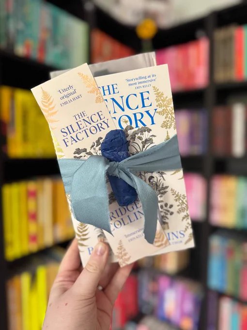 Hand holding "The Silence Factory" book with blue ribbon, against a blurred colorful bookshelf background. Text includes "Utterly original".