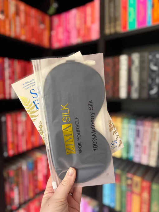 Hand holds a black sleep mask in a plastic pouch labeled "Zima Silk." Bookshelves with colorful books are in the blurred background.
