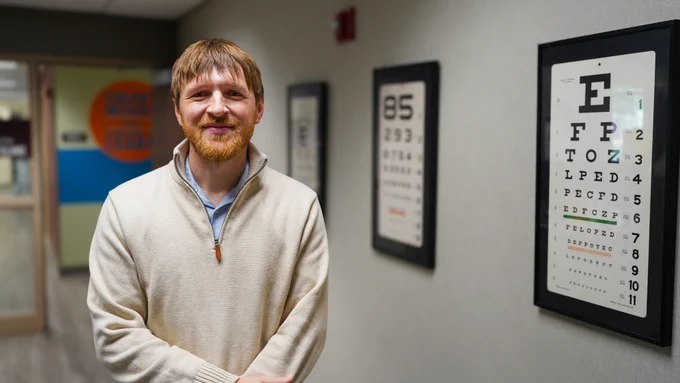 [Image description: Dr. Wiese is standing and smiling at the camera. He is in the entryway of the Low Vision Clinic. You can see various eye charts around him.]