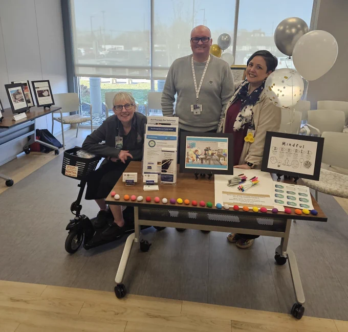 Three art therapy staff members pose behind a display table with colorful therapy materials. One person sits on a mobility scooter on the left, while two others stand behind the table, which features a "Mindful Doodle" poster, colorful clay balls, markers, and informational materials about Art Therapy for Veterans.