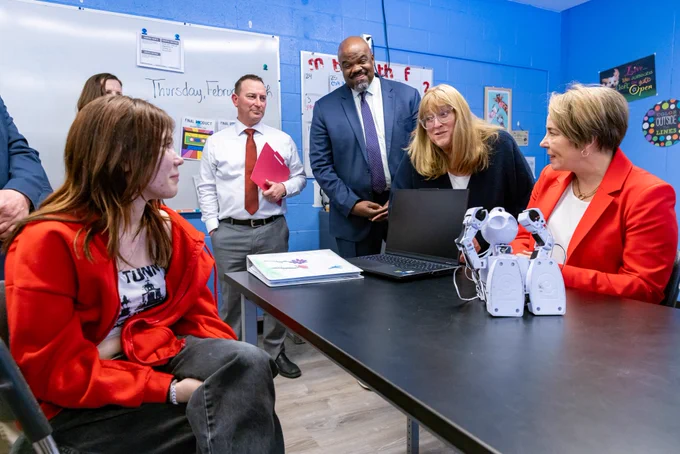 Governor Maura Healey, wearing a red blazer, sits at a table interacting with a small white humanoid robot and a laptop. Education Secretary Patrick Tutwiler, in a dark blue suit and tie, stands nearby smiling. They are joined by other adults and a student in a red sweatshirt, engaging in a classroom conversation surrounded by colorful student work and a whiteboard.