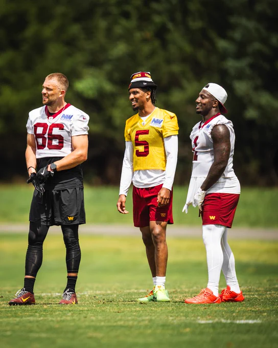 Photo of Zach Ertz, Jayden Daniels and Deebo Samuel Sr. outside during workouts. The three are standing together and smiling. Ertz and Samuel Sr. are wearing white practice jerseys, while Daniels is wearing a gold jersey. Ertz is waring black shorts, burgundy cleats and black football gloves. Daniels is wearing burgundy shorts, green cleats and a beanie hat. Samuel Sr. is wearing burgundy shorts, orange cleats, white football gloves and a white hat.