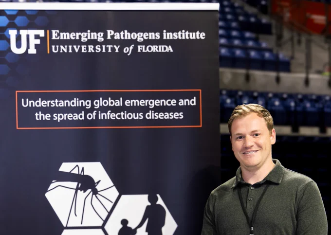 Scott Cohen smiles as he stands beside a vertical banner in the Stephen C. O'Connell Center during Emerging Pathogens Institute Research Day 2025. The vertical banner has the logo and the institute's mission: "Understanding global emergence and the spread of infectious diseases."
