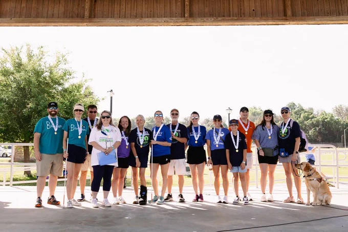 A group of 14 participants from the 2025 Jacksonville VisionWalk stand together at the finish line, wearing medals and smiling for the photo. The group includes men and women of various ages, some holding medals, and one participant is accompanied by a guide dog. They are standing under a pavilion with trees and clear skies in the background.
