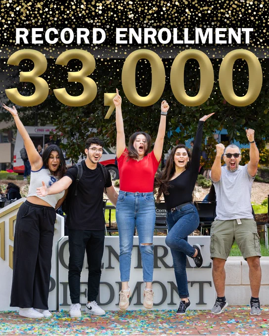 Five joyful students celebrating record enrollment of 33,000 at university, with two people lifting hands overhead in victory gesture, others cheering enthusiastically in front of the university sign.