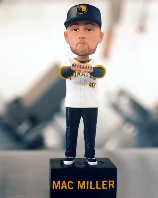 A bobblehead of Mac Miller. He's wear a black hat, Pirates home white jersey, dark blue jeans, and black and white shoes. The bobble is on a black stand with "Mac Miller" on the front in yellow letters.