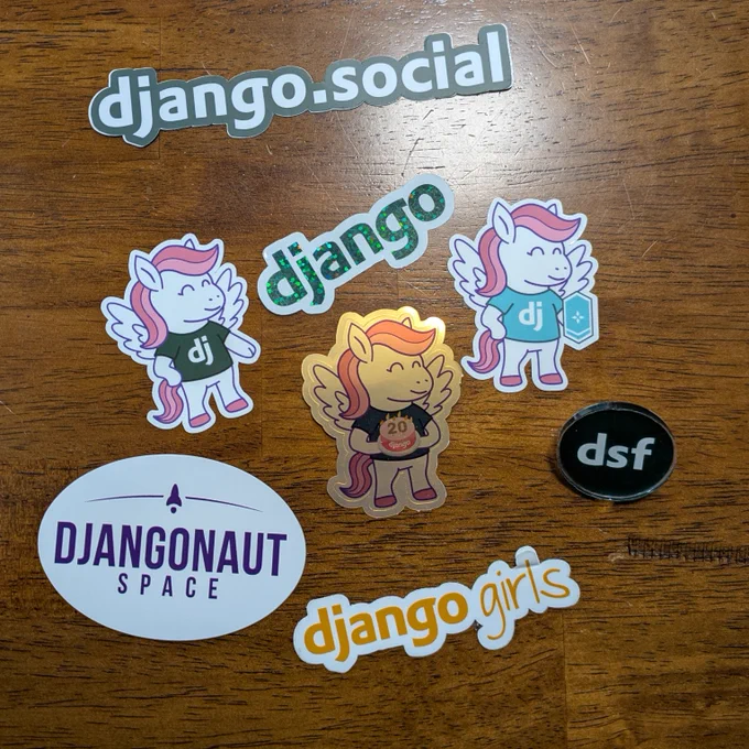 Close-up of Django stickers on a table - Django.social, Django pony with "dj" shirt, Django logo, Django pony with birthday cake, Django pony with shield, Djangonaut Space, DSF pin, Django Girls