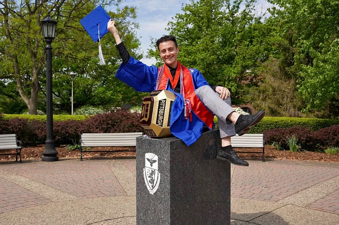 Braden Niles smiles while sitting inside of the Duquesne University Red Ring statue on campus while wearing his blue graduation gown. 