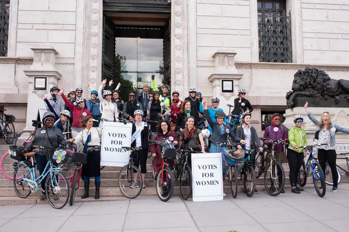 Group of individuals with bicycles gathered in front of a building, holding banners that read "Votes for Women.” 