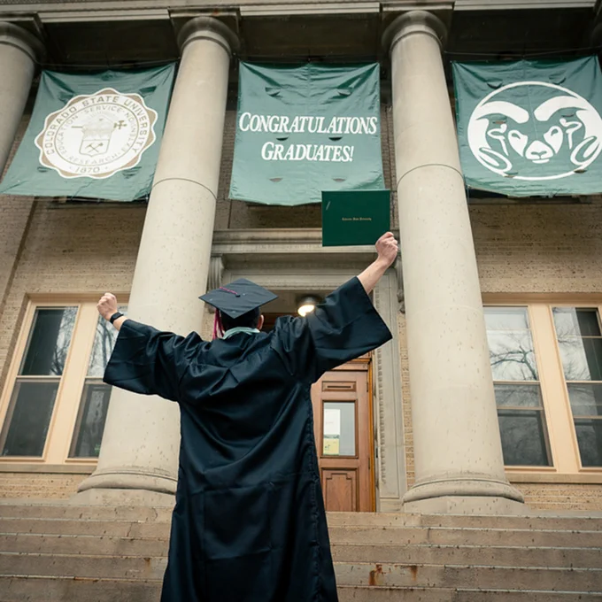 A C S U student dressed in commencement regalia triumphantly raises their arms high under a banner hanging from the Administration building that reads: Congratulations Graduates!"