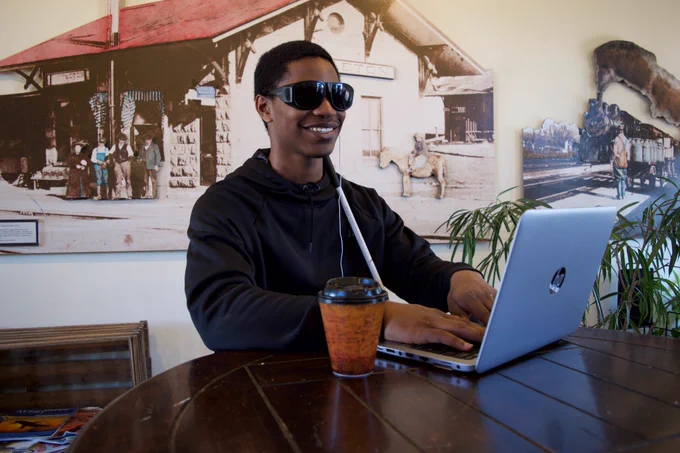 Photo of a blind man typing on a laptop. He is sitting by a round, brown table with his white cane by his side. A cup of coffee is next to the laptop.