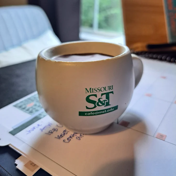 A white coffee cup stress ball with Missouri S&T's logo and cafe@mst.edu in green. It's sitting on a planner with notes for June, including "ATTW" at the top of the list.