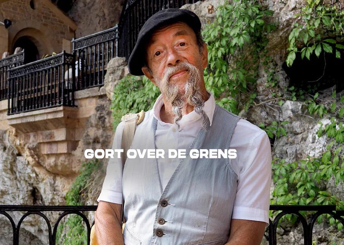Gort heeft een zwart baretje op en een grijs gilet boven op een wit shirt aan. Zijn grijze lange snor hangt in krullende slierten voorbij zijn kraag. Met een vredige glimlach kijkt hij vooruit.