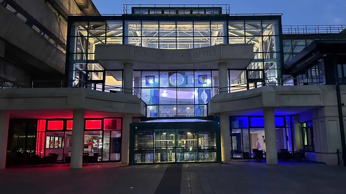 A photograph of The National Archives' building in early evening, lit up in red, white and blue.