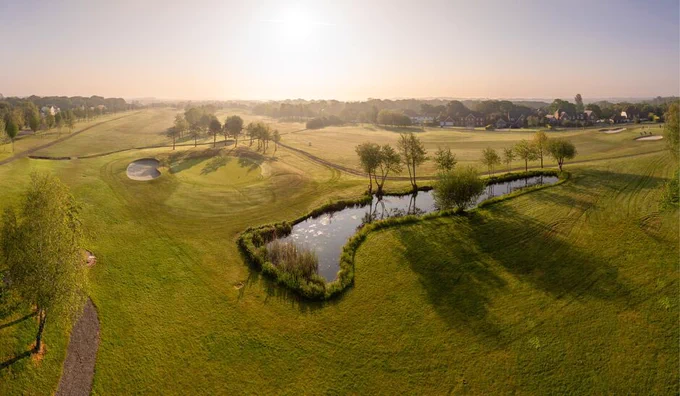 Aerial photograph of Skylark golf course taken at sunrise. In the middle of the course is a small pond surrounded by trees. The water is glistening. Trees surround the course. There is a bunker to the left of the pond.