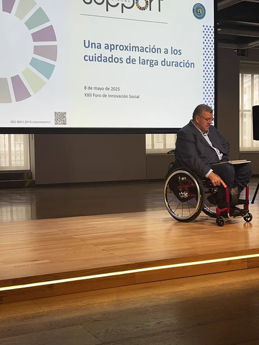 El dir de l’entitat sobre l’escenari durant la seva ponència. Al fons, una pantalla projecta la diapositiva “Una aproximación a los cuidados de larga duración”, amb el logotip de Support-Girona i una roda multicolor dels ODS. L’acte forma part del XXII Foro de Innovación Social, celebrat el 8 de maig de 2025.