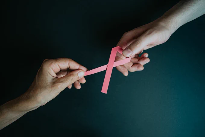 Two hands exchanging a pink ribbon symbolizing breast cancer awareness and support.