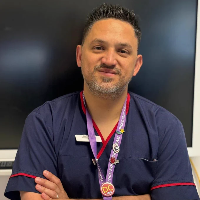Emanuele against a black background. He is wearing a nurse uniform with a purple lanyard around his neck.