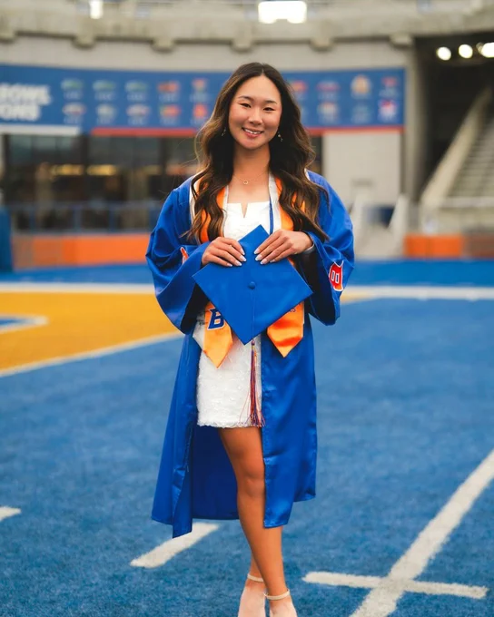 Boise State undergraduate student wearing graduation regalia smiling on the Blue Turf at Albertsons Stadium.