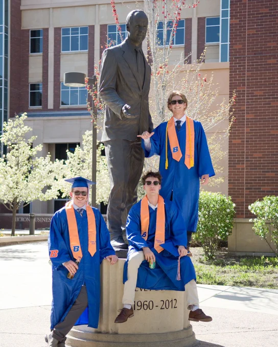 Three Boise State undergraduate students in graduation regalia posing next to bronze Steve Appleton statue.